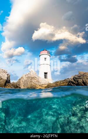 Split shot, over under shot. Half sky, half underwater. Defocused waves in the foreground with a lighthouse on a rocky coast. Stock Photo