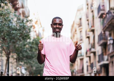 horizontal portrait of a handsome young african man standing in the middle of a street. He is doing positive gesture with his fingers Stock Photo