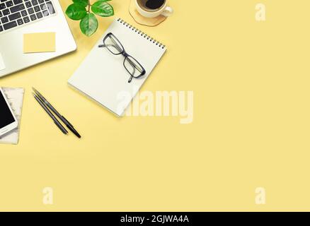 Yellow office desk table with laptop computer, green plant, pens, glasses, smurtphone and a cup of coffee. Top view with copy space, flat lay. Stock Photo