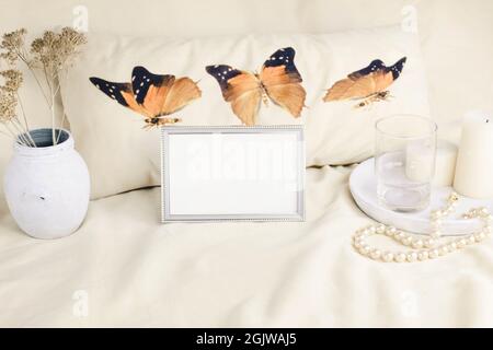 Portrait metal frame mockup on the background of a pillow with butterflies, a vase, white candles and a necklace of pearls, an empty glass on a white Stock Photo