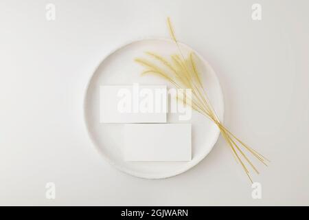 Mockup of business cards on a white vintage plate with dry plant on gray table background in a elegant minimalist style. Top view. Stock Photo