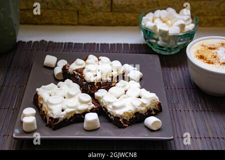 A shot of marshmallow cake pieces on a tray, a bowl of marshmallows, and a cup of cappuccino on a wooden table Stock Photo