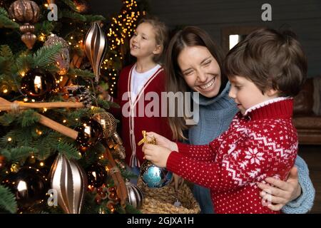 Laughing young mother enjoying decorating Christmas tree with small children. Stock Photo