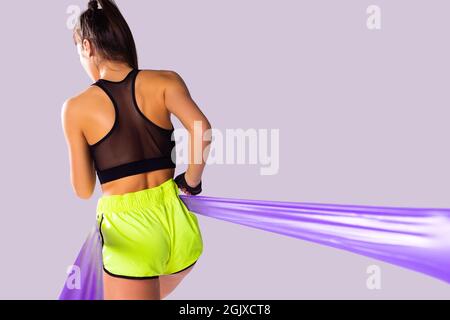 Back photo of a slim athletic young woman in sportive bodysuit doing strength training using resistance band. Stock Photo
