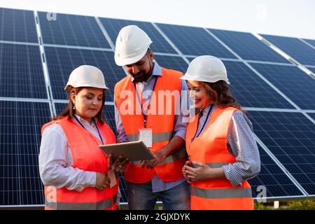 The happy group of engineers standing against the backdrop of solar panels. They communicate lively and look at the tablet Stock Photo