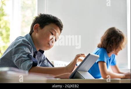 Concentrated Asian schoolboy using tablet computer device in classroom. Stock Photo