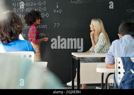 Afro American schoolkid answering task to teacher near chalkboard in classroom. Stock Photo