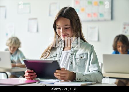Happy smiling schoolgirl reading task from tablet computer device in classroom. Stock Photo