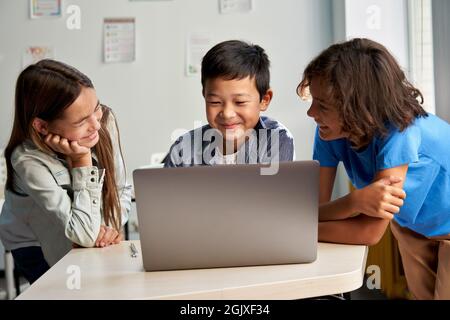 Three diverse schoolmates watching video together on laptop in classroom. Stock Photo