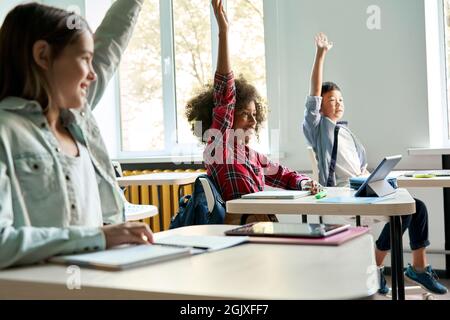 Diverse schoolkids sitting at desk raising hands answering at lesson. Stock Photo