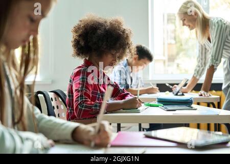 Diverse classmates doing task at lesson studying in classroom with teacher. Stock Photo