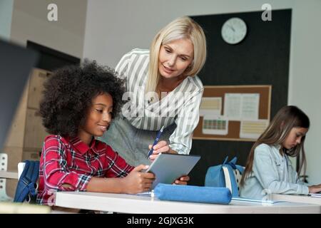 Happy Afro American schoolkid listening to teacher holding tablet in classroom. Stock Photo