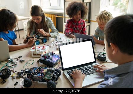 Diverse schoolkids at stem class building robot using pc with white screen. Stock Photo