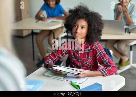 Happy Afro American schoolgirl looking at teacher holding tablet in classroom. Stock Photo
