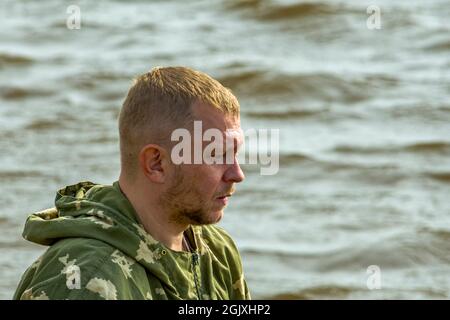 Unshaven fisherman against background of waves on river looks forward. Side view. In nature, outdoors horizontal shot. Stock Photo