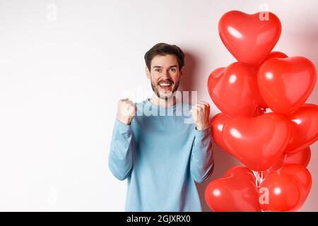 Portrait of happy guy celebratiing lovers day, standing near valentines red heart balloon and cheering, making yes gesture and smiling at camera Stock Photo