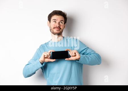 E-commerce and shopping concept. Smiling young man showing mobile screen and dreaming about something, looking at upper right corner nostalgic Stock Photo