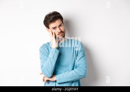 Sad young man looking frustrated, touching temple of head and frowning uneasy, standing against white background Stock Photo