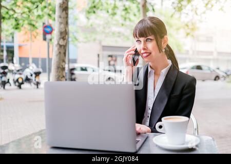 Portrait of beautiful businesswoman using laptop sitting in coffee shop outdoors and using smartphone Stock Photo