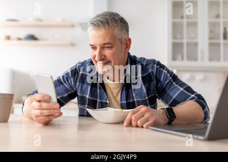 Happy mature man using smartphone while having breakfast in kitchen table at home, copy space Stock Photo