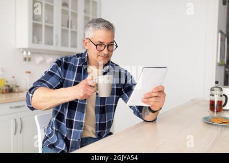 Morning routine. Senior man reading his notepad while sitting at kitchen table and drinking coffee, free space Stock Photo