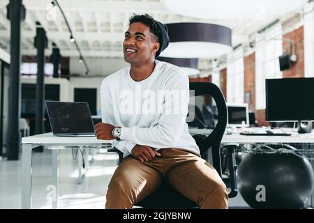 Happy software developer smiling in a modern office. Confident young businessman looking away while sitting alone. Young businessman taking a break wh Stock Photo