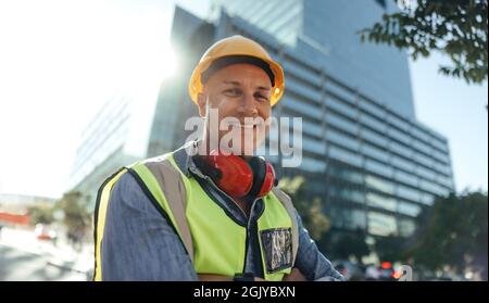 Cheerful construction worker smiling at the camera while standing with his arms crossed in the city. Mid-adult blue collar worker standing in front of Stock Photo