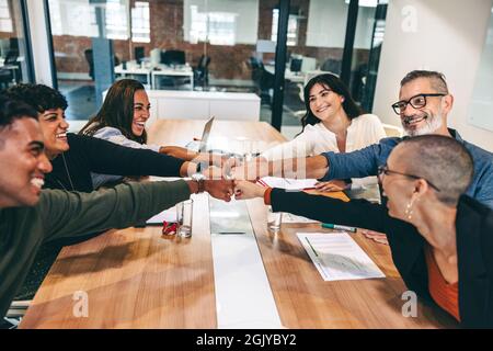 Team of happy businesspeople bringing their fists together while sitting in a boardroom. Diverse group of colleagues smiling cheerfully during a meeti Stock Photo