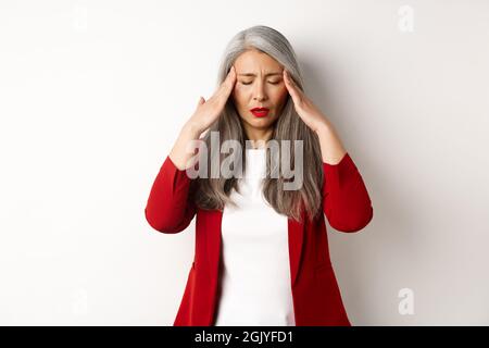 Tired senior businesswoman in red blazer having headache, touching head and feel sick, standing with migraine against white background Stock Photo