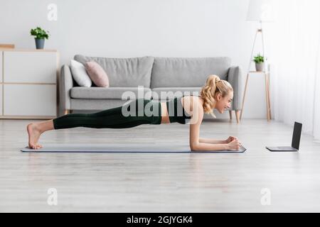 Sad european pretty millennial blonde lady doing plank and watch at laptop with workout video lesson at home Stock Photo