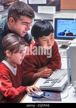 Early 1990s computer school lessons study Junior 2nd grade blond schoolgirl 7-9 years with male teacher and 7 years old oriental Korean boy studying enthusiastically at an older style computer terminal with mouse and UV protection screen in junior school classroom In the 1990s Stock Photo