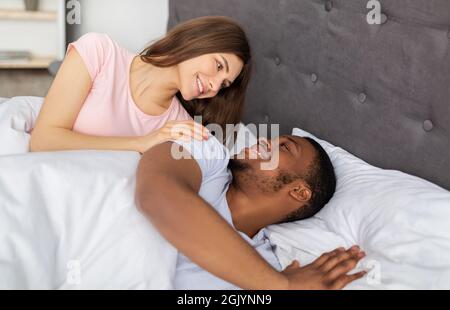 Loving married diverse couple relaxing, looking at each other in bed at home, feeling carefree and happy in morning Stock Photo