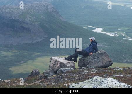 Female hiker in blue jacket overlooking remote arctic valley on a cloudy day of arctic summer. Hiking in Sarek National Park, Sweden, with Rapa River Stock Photo