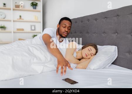 Jealous Afro man checking his sleeping girlfriend's mobile phone in bed at home. Marriage problems concept Stock Photo