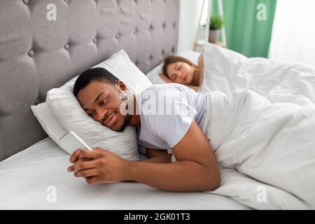Young black guy lying on bed and using mobile phone to text his lover while his wife is asleep. Relationship infidelity Stock Photo