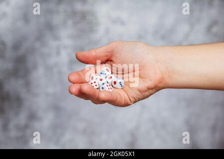 A man's right hand holding several white dice with red and blue dots on a textured gray background. Stock Photo