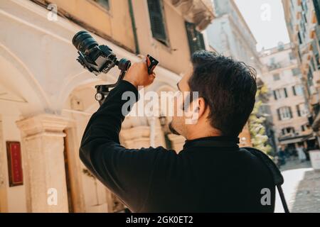 Male cameraman shoots urban architecture and building facades with steadicam on mirrorless camera. Greece or Italy background. Stock Photo