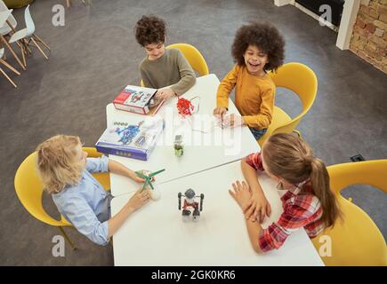 Interesting classes. High angle view of happy diverse kids sitting at the table, examining technical toys full of details, spending time at Stock Photo