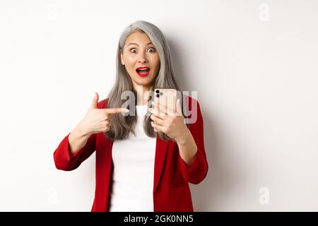 Amazed asian lady in red blazer pointing finger at smartphone, staring impressed at camera, showing something online, white background Stock Photo