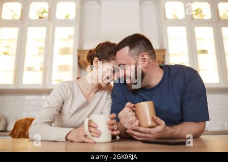 Happy affectionate young Caucasian couple leaning on kitchen table and laughing while drinking coffee together Stock Photo