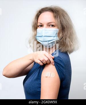 Vaccinated young woman showing shoulder with plaster, pretty person in mask after getting COVID-19 vaccine. Concept of people coronavirus vaccination, Stock Photo