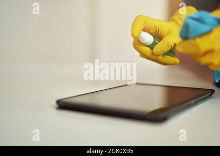 Close up shot of cleaning lady in rubber gloves using antibacterial sanitizer spray and cloth while disinfecting the screen of digital tablet pc Stock Photo