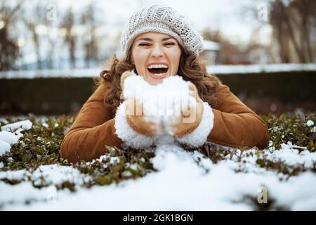 smiling elegant 40 years old woman with mittens in a knitted hat and sheepskin coat playing outdoors in the city park in winter. Stock Photo