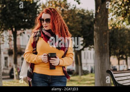 Portrait of young red-haired woman in yellow sweater and sunglasses. She holds reusable coffee mug in her hands and walks through park on sunny autumn Stock Photo