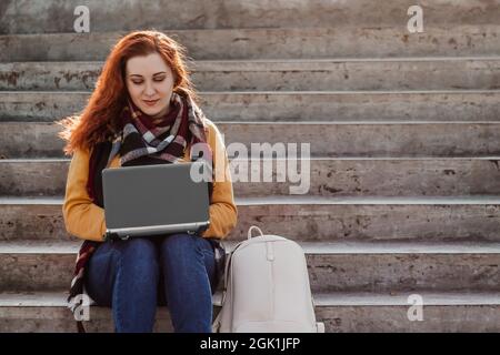 Young red-haired woman sits on stairs and communicates on Internet using laptop. Modern businesswoman freelancer is working outdoor. Hipster with gadg Stock Photo