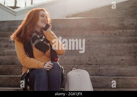 Young red-haired woman sitting on stairsand talking on phone. Lady in yellow sweater uses smartphone on sunny day. Lifestyle, copy space. Stock Photo