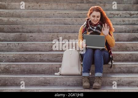Young red-haired woman sits on stairs and communicates on Internet using laptop. Modern businesswoman freelancer is working outdoor. Hipster with gadg Stock Photo