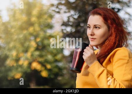Young red-haired woman drinks coffee from reusable thermo mug. Enjoy autumn sunny day in nature. Eco-friendly lifestyle. Stock Photo