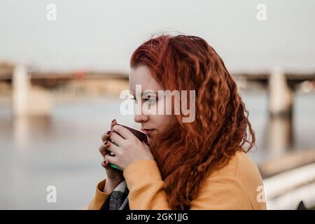 Portrait of young red-haired woman against background of city river and bridge. She drinks coffee from paper cup and enjoys autumn view. Copy space. Stock Photo