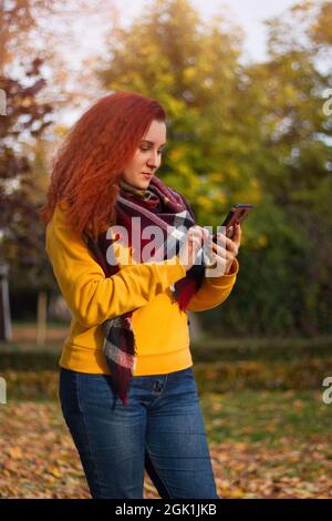 Red-haired curly woman uses modern smartphone. She walks in autumn park. Chatting and messaging on the Internet. Stock Photo